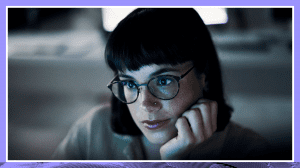 Woman with a bob haircut and glasses sits with her hand on her chin looking at a computer screen. Photo has darker purple and blue tones.