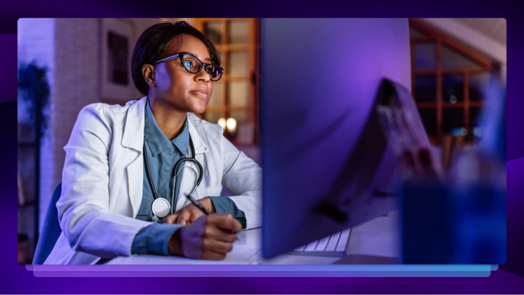 A doctor writes on paper while looking at a computer monitor with a stethoscope around her neck.