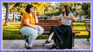 Photograph of two women sitting on a park bench. They appear to be using sign language they’re using their hands to communicate with one another. Woman on the left is wearing jeans, a yellow shirt sneakers, glasses. Woman on the right is wearing a black and white polkadot blouse, black shirt, sneakers, glasses. Trees in the background are out of focus and glowing with a soft golden light.