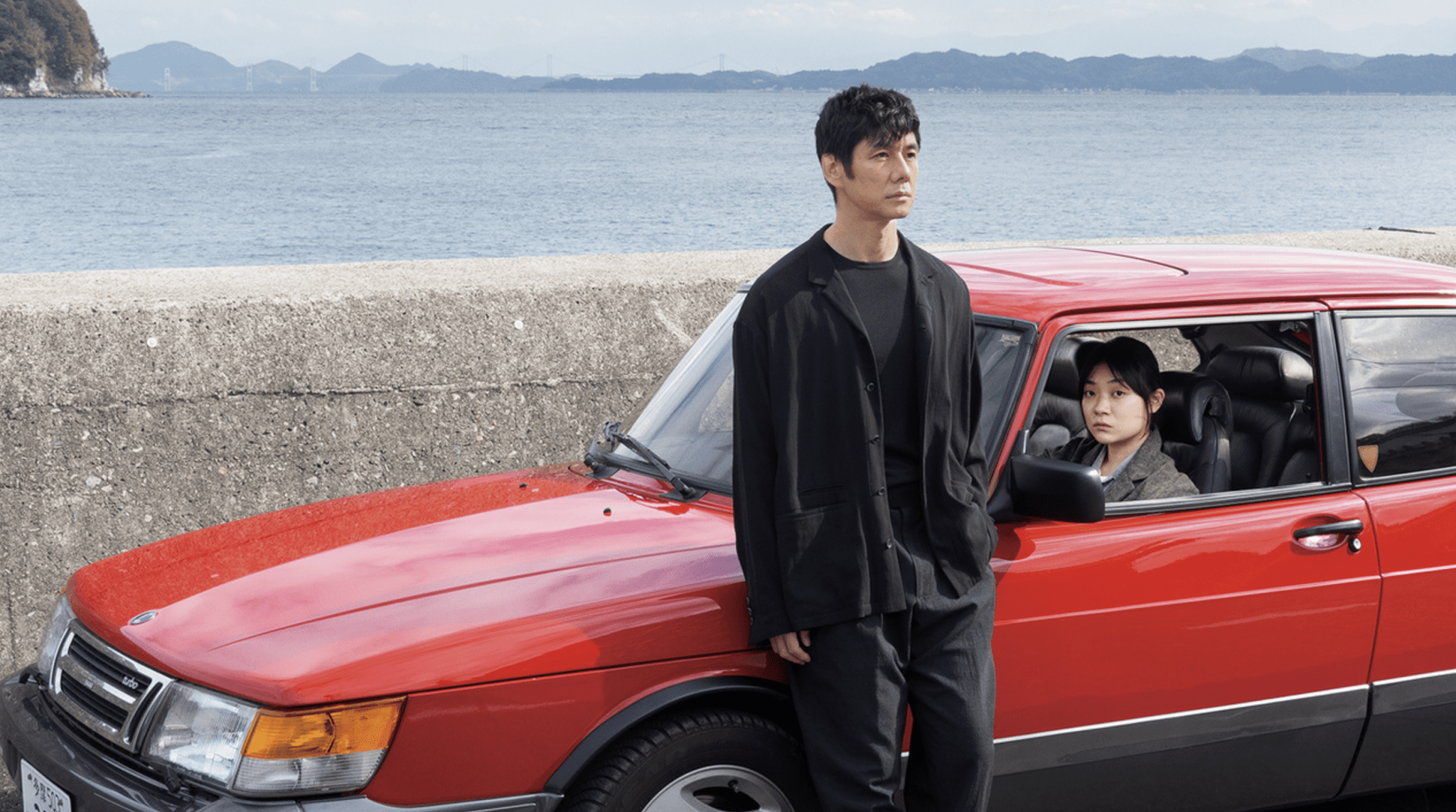 Hidetoshi Nishijima in Drive My Car leans against a red Saab staring pensively into the distance. A female driver sits in the front seat looking at the camera. The background is of the Japanese coastline with faint mountains in the distance.
