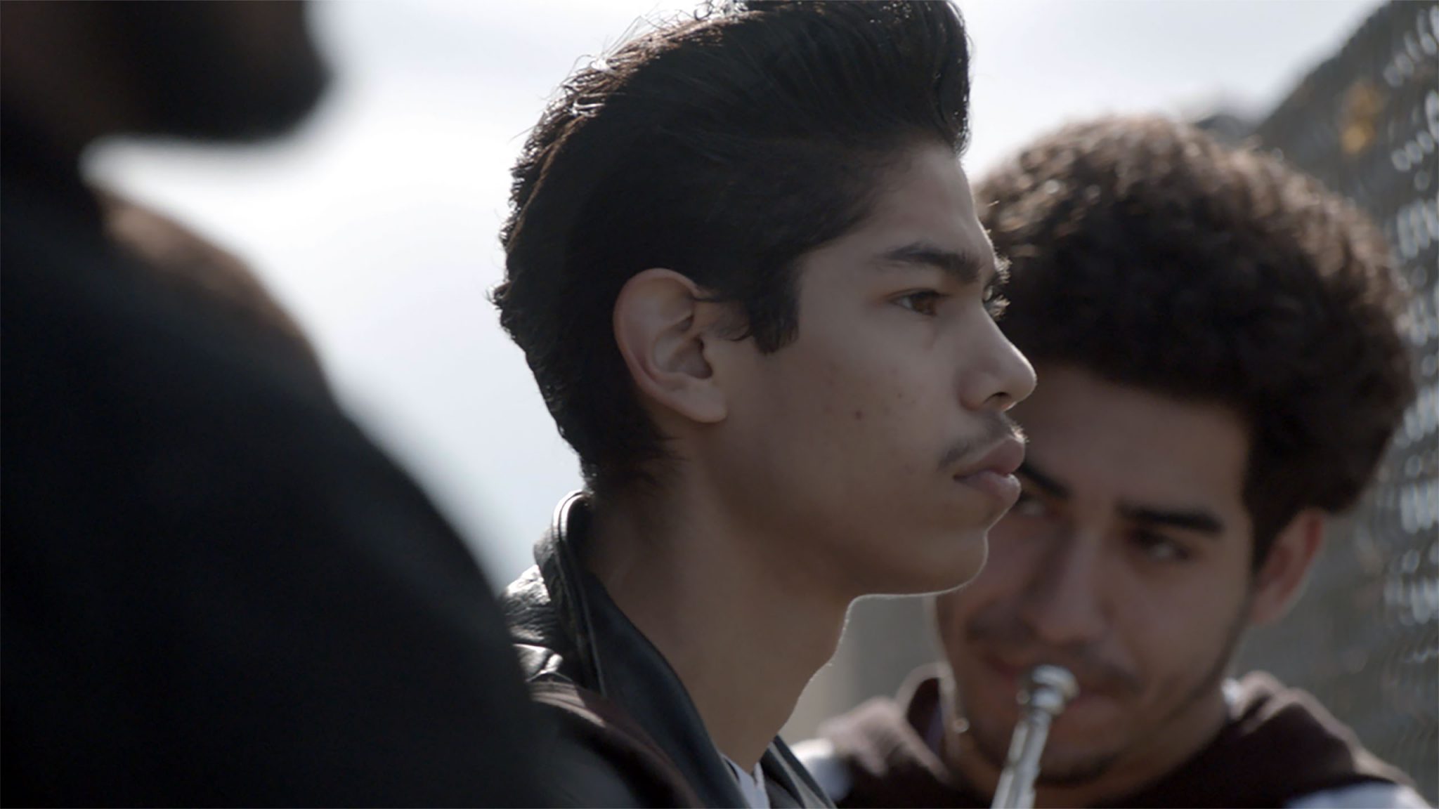 Photo still from ‘The Tuba Thieves,’ showing two high school boys outside at what could be a band practice. One boy is out of focus with a brass instrument held up to his lips. We see the other boy’s side profile and he’s gazing out of frame.