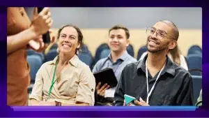 A man and a woman are sitting, smiling, and taking notes while listening to a speaker give a presentation.