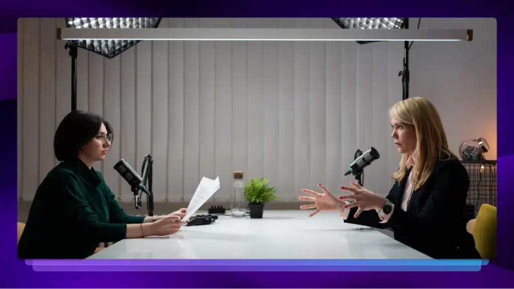 Two women sit across from each other at a table with microphones, conducting a recorded interview.