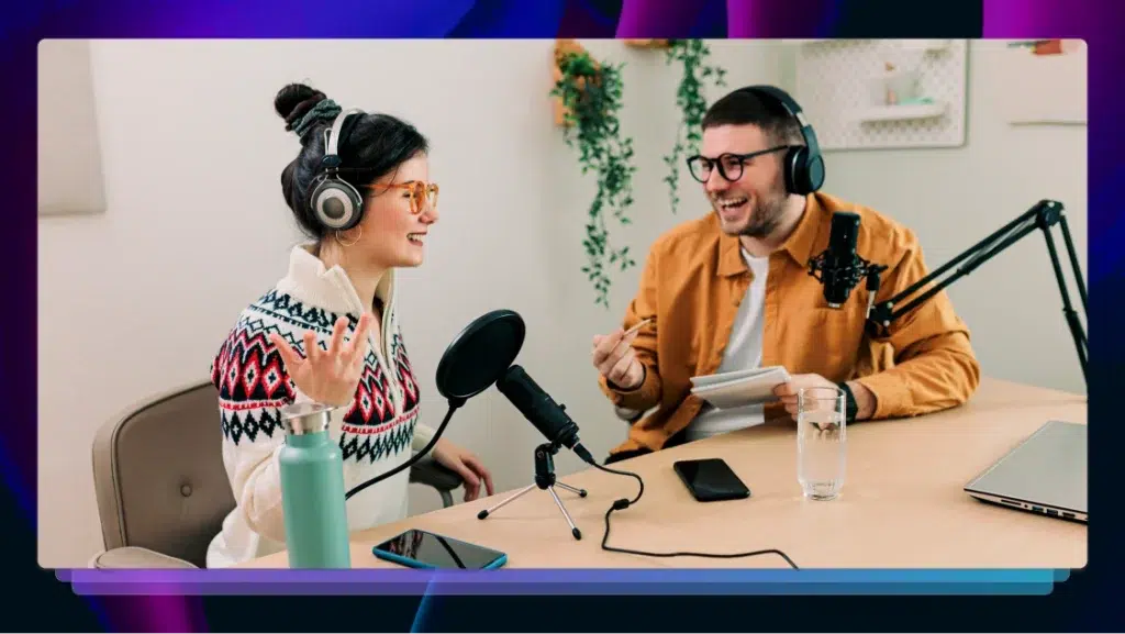 A podcast host interviews a guest at a wooden table with microphones in front of them. They are both smiling and laughing.