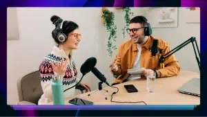 A podcast host interviews a guest at a wooden table with microphones in front of them. They are both smiling and laughing.